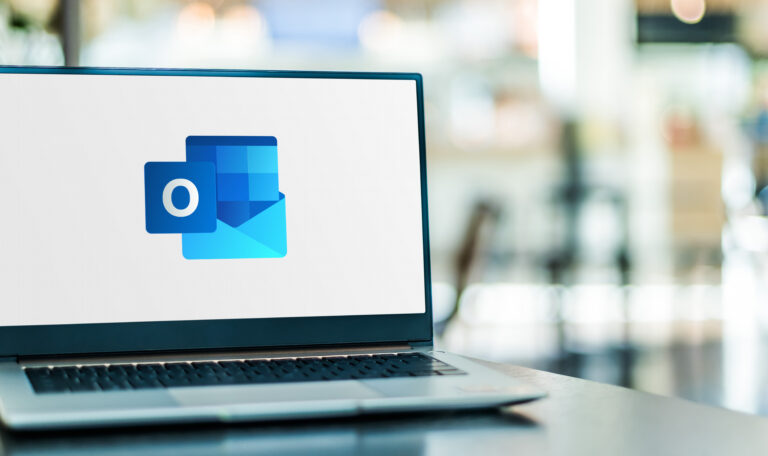 A laptop displaying the Microsoft Outlook logo on a desk in a modern office, with a softly blurred background.