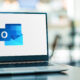 A laptop displaying the Microsoft Outlook logo on a desk in a modern office, with a softly blurred background.