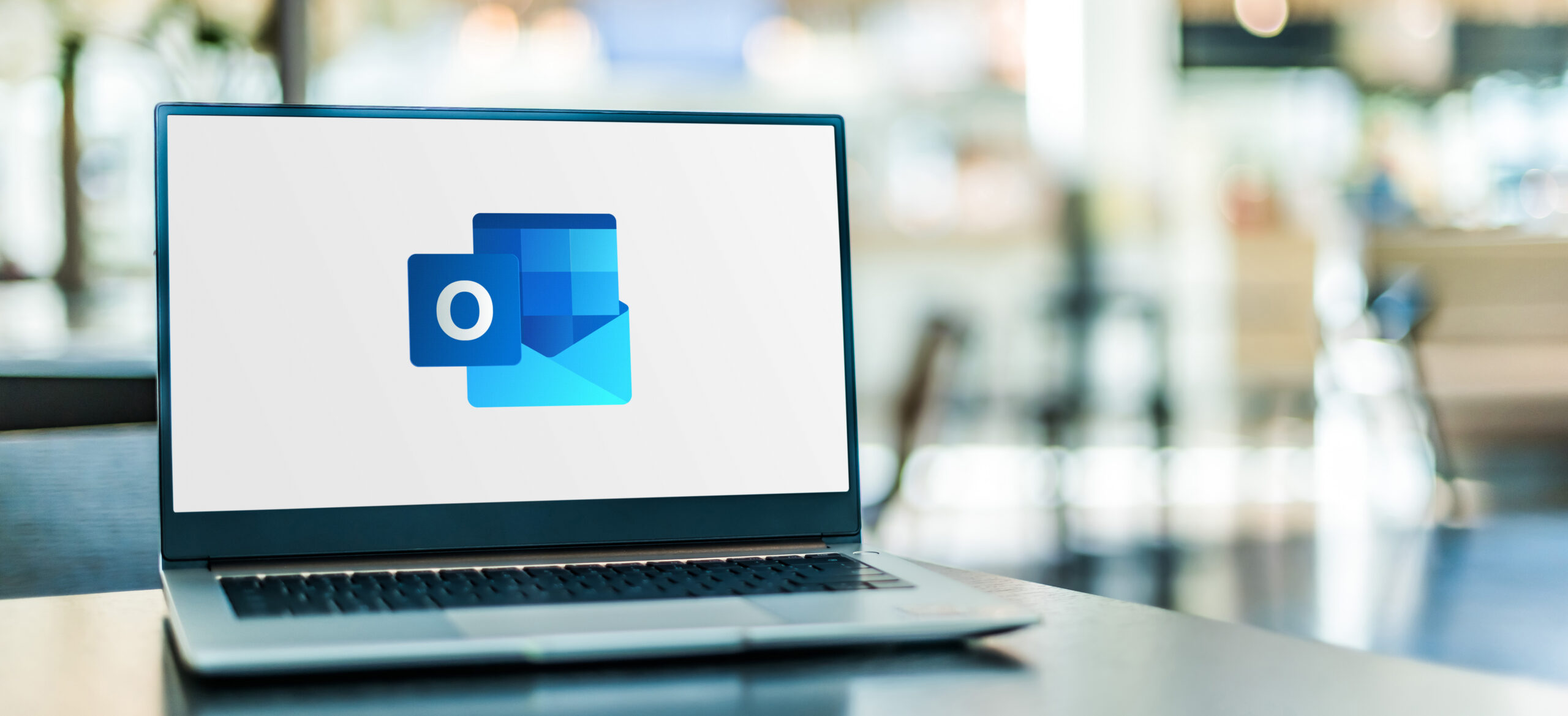 A laptop displaying the Microsoft Outlook logo on a desk in a modern office, with a softly blurred background.