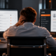 Stressed account manager seated between two monitors, with an email inbox on one screen and a separate CRM dashboard on the other, surrounded by paperwork and office supplies in a dimly lit office with charcoal grey tones and warm orange accents, illustrating the challenges of CRM adoption and disconnected workflows.